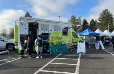 A green and white Embold Credit Union mobile branch is parked in a lot. People stand in line at the truck, and nearby are blue and white tents with tables and displays. Trees and a clear sky are in the background. EMBOLD Credit Union