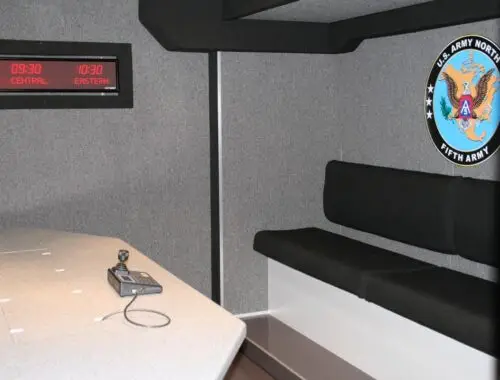 A meeting room with a gray interior, a wall-mounted US Army North Fifth Army emblem, a digital clock display showing multiple time zones, a table, and a small electronic device with a cord. Army North 1