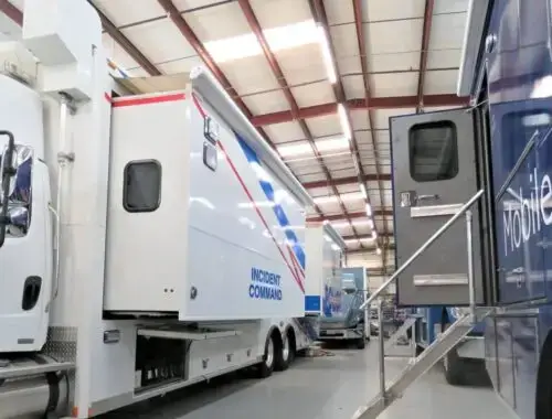 Two large specialized vehicles are parked inside a warehouse. One is labeled “Incident Command” and the other has partial text Mobile Bra. Stairs lead up to the blue vehicle’s entrance. The ceiling is made of metal beams.