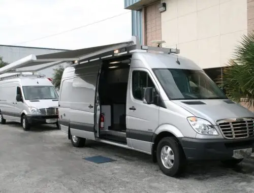 Two silver vans are parked outside a building, each with a large extended side awning. The front van has its side door open, revealing some interior equipment. The scene appears to be in an industrial area. UAV Sprinter Light Duty