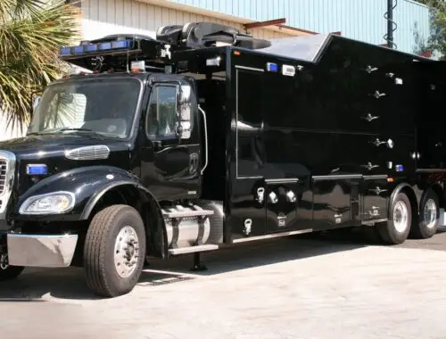 A large black specialized truck with multiple compartments and equipment, parked outside near a building and palm tree; the truck has official markings on the side and visible emergency lights on top. Army North 1