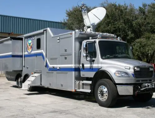 A large, silver mobile command center truck with blue stripes, a satellite dish mounted on top, and multiple compartments is parked on pavement near a building and trees under a clear sky. Nevada National Guard EHD.
