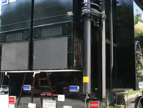 A large black mobile command vehicle with multiple vents, lights, and a telescoping antenna is parked outdoors. The back panel displays a license plate and equipment compartments. Army North 1