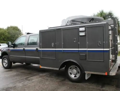 A gray utility truck with multiple storage compartments on the side is parked on a wet street. The truck has a blue stripe and a mounted satellite dish on its roof. Trees and another vehicle are in the background. Nevada National Guard RIV
