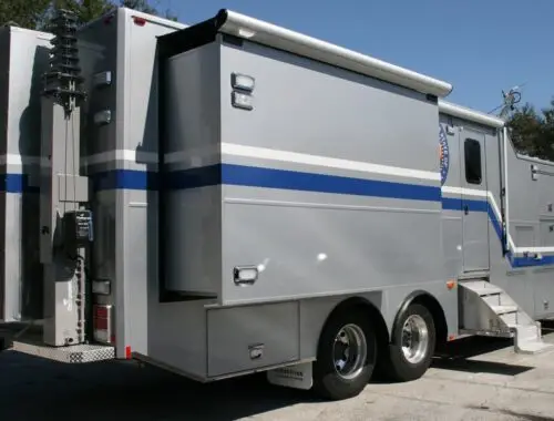 A large gray mobile command center vehicle with blue and white stripes, dual rear wheels, a side door with steps, equipment mounted on the rear, and an extended awning on the side, parked outdoors.