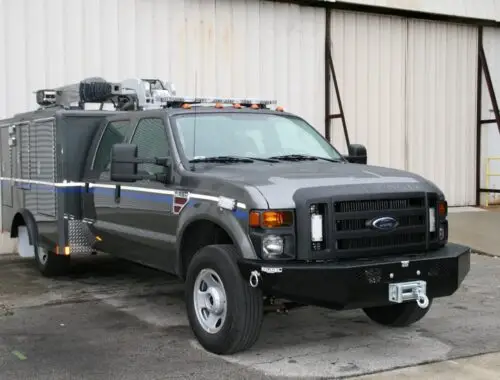 A large, armored Ford truck is parked on a paved surface in front of a beige metal building with a roll-up door. The truck features heavy-duty bumpers, protective plating, and specialized equipment mounted on the roof. Nevada National Guard RIV