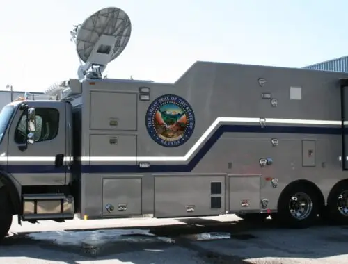 A large, gray mobile command vehicle with a satellite dish on top and the seal of the State of Florida on the side is parked outside an industrial building.