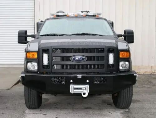 A black armored vehicle, viewed from the front, is parked on an asphalt surface near a beige industrial building with a white roll-up door. The vehicle has large side mirrors and visible Ford branding on the grille. Nevada National Guard RIV