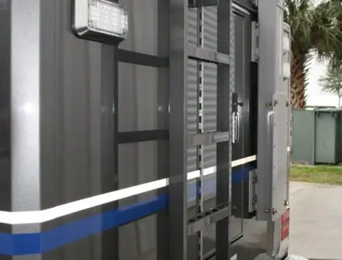 A close-up view of the back of a dark-colored truck or emergency vehicle with a mounted black metal ladder, various lights, and a blue stripe; a palm tree and some outdoor structures are visible in the background.