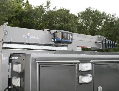 A close-up view of a Floatograph telescoping mast mounted on top of a dark utility vehicle, with several lights and equipment cabinets visible below, and trees in the background.