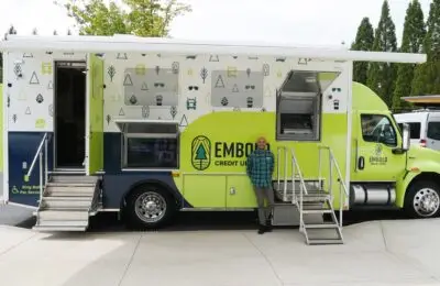 A man stands smiling in front of a green and white Embold Credit Union mobile banking truck with stairs leading to two entrances on the side, parked outdoors near trees and pavement. EMBOLD Credit Union