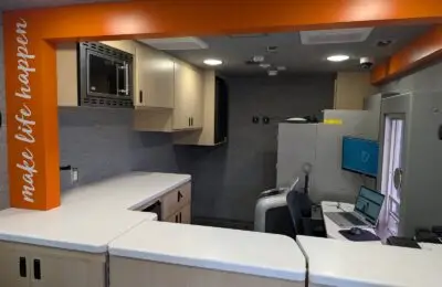 A modern office space with light wood cabinets, a microwave, a computer workstation with two monitors, a printer, and an orange beam with the words “make life happen” on it.