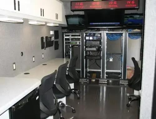 Interior of a mobile broadcast control room with several office chairs, workstations with countertops, wall-mounted equipment racks, monitors displaying different time zones, and overhead storage cabinets.