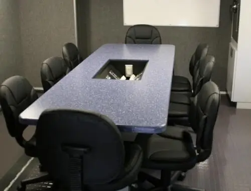 A conference room with a rectangular table surrounded by eight black office chairs. The table has a recessed center holding markers. A blank whiteboard is mounted on a gray wall in the background.