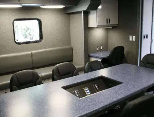 A modern conference room with a long blue table, black chairs, a built-in control panel, gray bench seating, wall cabinets, a small desk area, and a window. The room has gray walls and bright lighting.