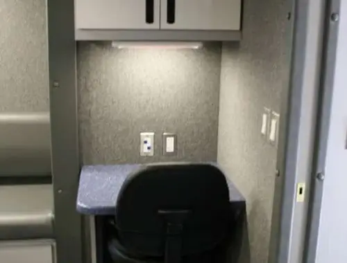 A small workstation with a chair, a blue speckled desk, two power outlets, a light fixture, and a set of white cabinets mounted above the desk against a gray wall.