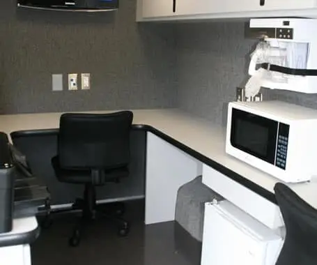 A small office space with a desk, office chair, wall-mounted television displaying a weather map, microwave, mini fridge, cabinets, and electrical outlets. The setting appears organized and functional.