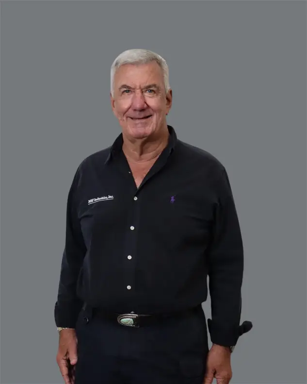An older man with short gray hair stands facing the camera, wearing a dark MBF Industries company button-up shirt with a badge on the chest and dark pants, set against a plain gray background.