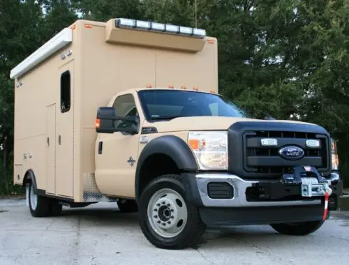A tan Ford truck with a large, boxy utility body and emergency lights is parked on pavement near trees. The truck has a front winch and black bumper, and appears to be a specialized emergency or service vehicle.