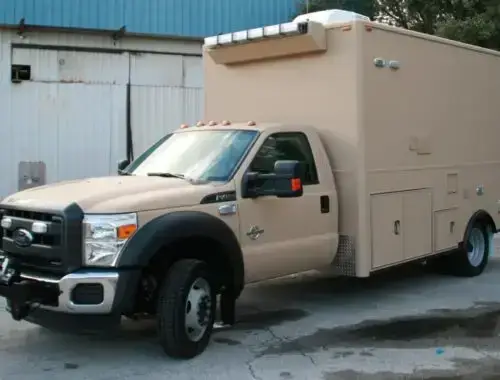 A tan utility truck with a large enclosed cargo area is parked on a paved surface near a metal building with a blue trim and some trees in the background.