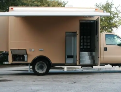 A large tan utility truck is parked with its side door open, an extended awning, and a loading ramp deployed at the rear. The truck has metal steps leading inside and utility compartments along the side.