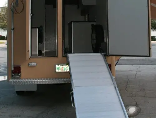 A tan utility vehicle with its back doors open and a metal ramp extended to the ground. The interior of the vehicle is partitioned and visible, showing some equipment and a spare tire.