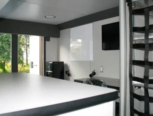 Interior of a small modern room with a white countertop in the foreground, a desk and chair, a wall-mounted TV, a whiteboard, and a windowed door open to a view of trees outside.