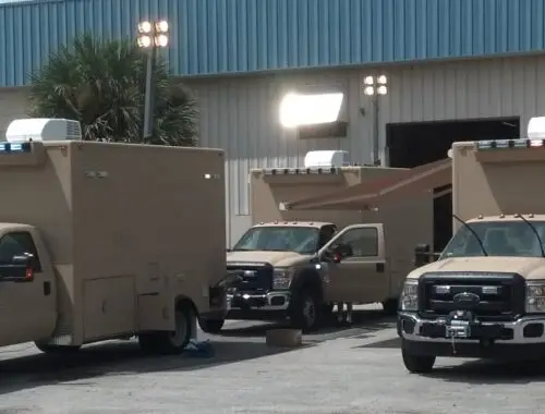 Four tan armored trucks with blue and white lights are parked outside a large industrial building with open roll-up doors. Some doors on the trucks are open, and overhead lights are mounted on the building.