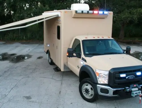 A tan Ford emergency response vehicle with blue and red lights on the roof is parked on a paved surface. An awning extends from the side. Trees are visible in the background.