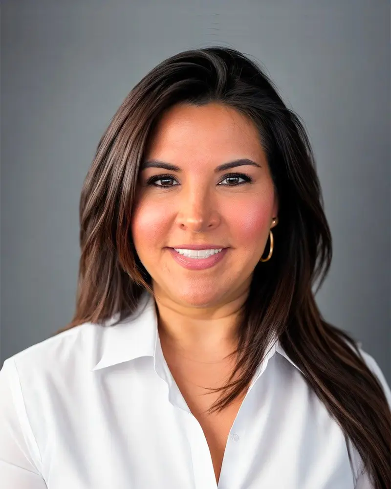 A woman with long brown hair, wearing a white collared shirt and gold hoop earrings, smiles at the camera against a plain gray background, representing MBF Industries company.
