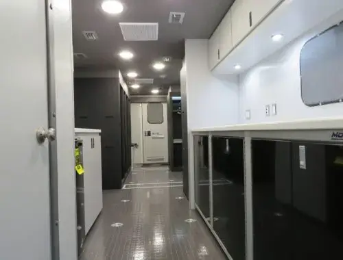 Interior view of a mobile command center or specialized vehicle, showing clean counters, storage cabinets, overhead lighting, and secure doors, with a non-slip floor and various electrical outlets.