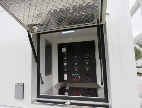 A rectangular outdoor electrical control panel with various switches, buttons, and outlets is mounted inside a metal compartment. The compartment has a diamond-plate lid held open, and a small light is installed at the top.