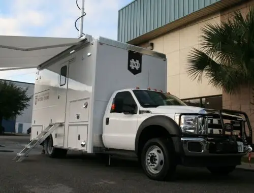 A large white mobile command vehicle with the Notre Dame logo is parked outside a building. The vehicle has an awning extended, several compartments, and a door accessed by steps. A palm tree is visible nearby.