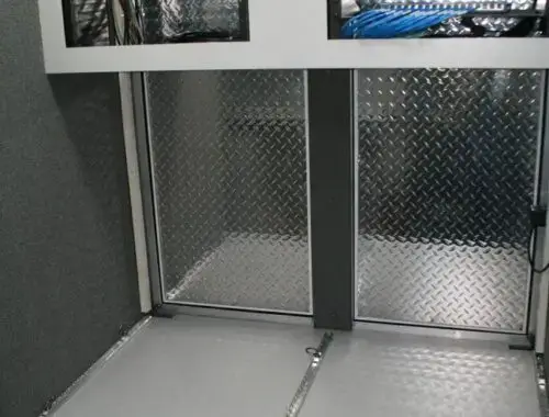 A metal storage compartment with diamond plate doors and panels, open shelving with exposed wiring and cables, and closed lower cabinets with black handles. The area appears clean and organized.