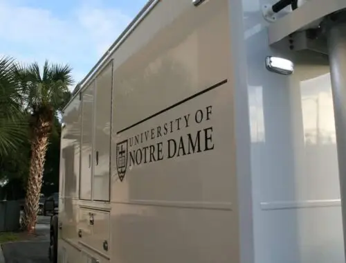 A white vehicle with “University of Notre Dame” and a university crest logo printed on the side, parked near palm trees under a partly cloudy sky.
