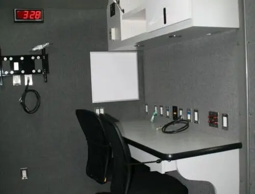 A small office space with a desk, two black chairs, overhead white cabinets, multiple wall outlets, a digital clock displaying 3:28, and various cables hanging on the wall.