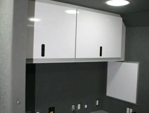 A small office workspace with a gray desk, two black chairs, white overhead cabinets, a whiteboard on the wall, and multiple electrical outlets and ports above the desk, all within a gray, carpeted room.