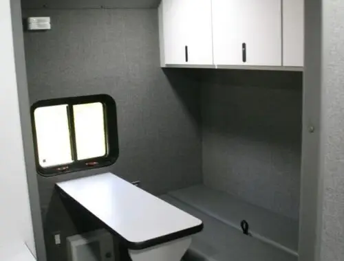 A compact, modern RV interior with gray walls, a small window, overhead white cabinets, a long bench seat with gray upholstery, and a narrow table attached to the floor. Ceiling lights are turned on.
