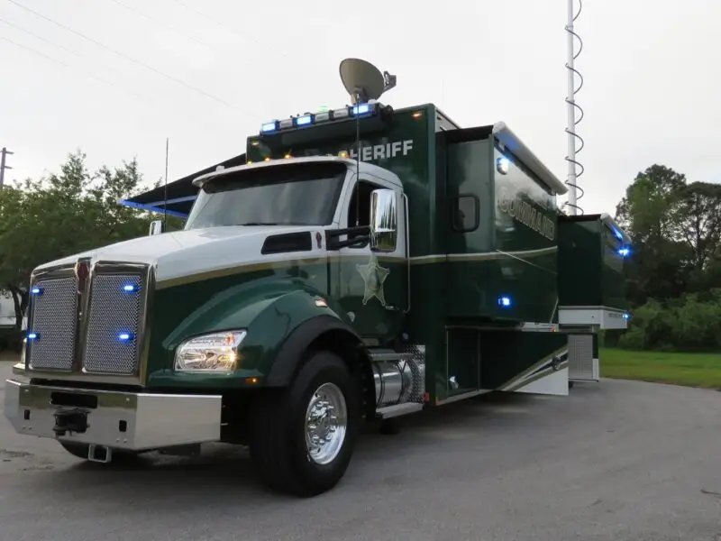 A large green Volusia County Sheriffs Office Public Safety Vehicle with flashing blue lights is parked on a paved area near trees, featuring antennas and communication equipment on the roof.