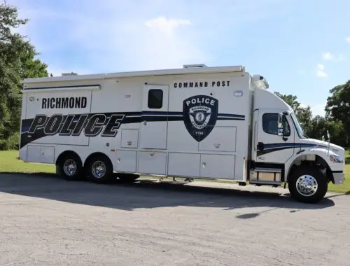 A large white Richmond Police command post vehicle is parked on a paved surface with grass and trees in the background under a clear blue sky.