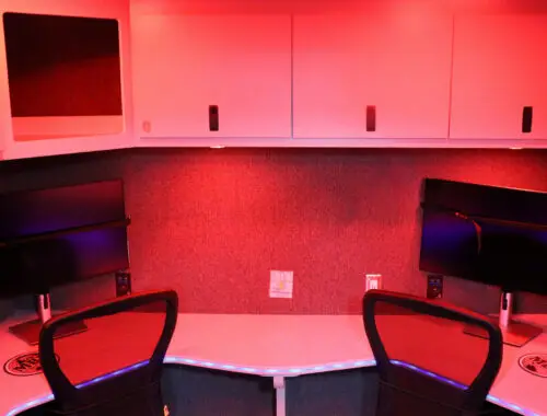 Two black office chairs sit at a curved white desk with two large computer monitors on a gray wall. There are white cabinets above the desk, and the room is lit with a red light.