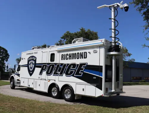 A large white Richmond Police command vehicle is parked on a paved area near grass and trees. The vehicle has a raised surveillance camera mast on the roof and police markings on the side.