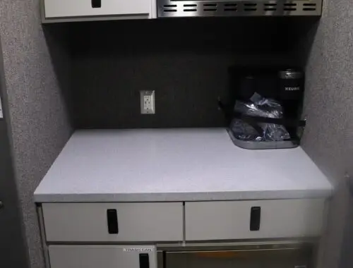 A small kitchenette with a microwave oven, a cabinet, a Keurig coffee maker, two drawers, a mini-fridge, and a gray countertop against a gray wall.