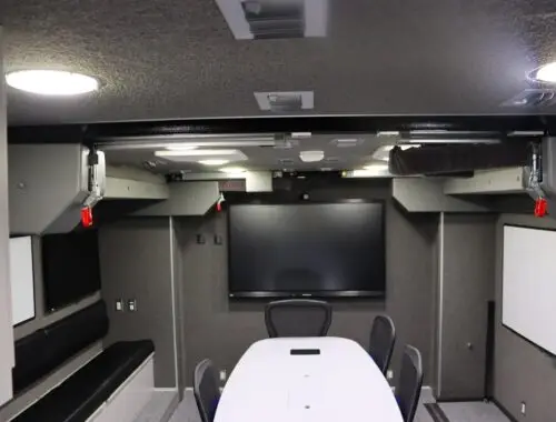 A modern conference room with a long oval table, five chairs, a large flat-screen monitor on the wall, whiteboards on both sides, and built-in bench seating along one wall. Ceiling lighting illuminates the room.