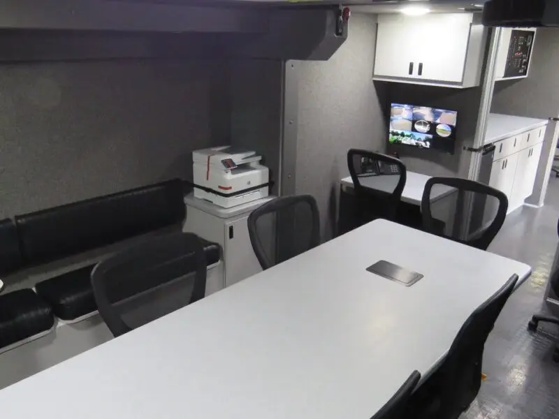 A modern, compact mobile office with a long table, office chairs, bench seating, a printer, wall-mounted monitors displaying surveillance footage, and built-in white cabinets. The space is well-lit and organized.